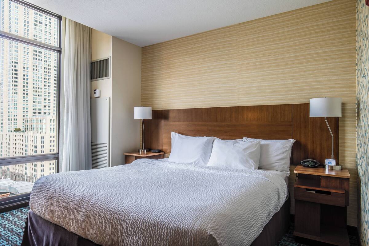 Hotellihuone, jossa on suuri sänky, kaksi yöpöytää, puinen sängynpääty ja suuri ikkuna, josta avautuu näkymä kaupunkimaisemaan.