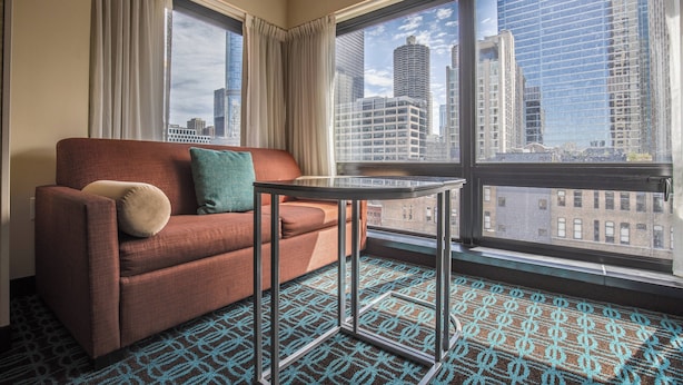 A hotel room with a red sofa, a small table, and a view of city buildings through large windows. - Fairfield Inn & Suites Chicago Downtown/River North (Chicago)