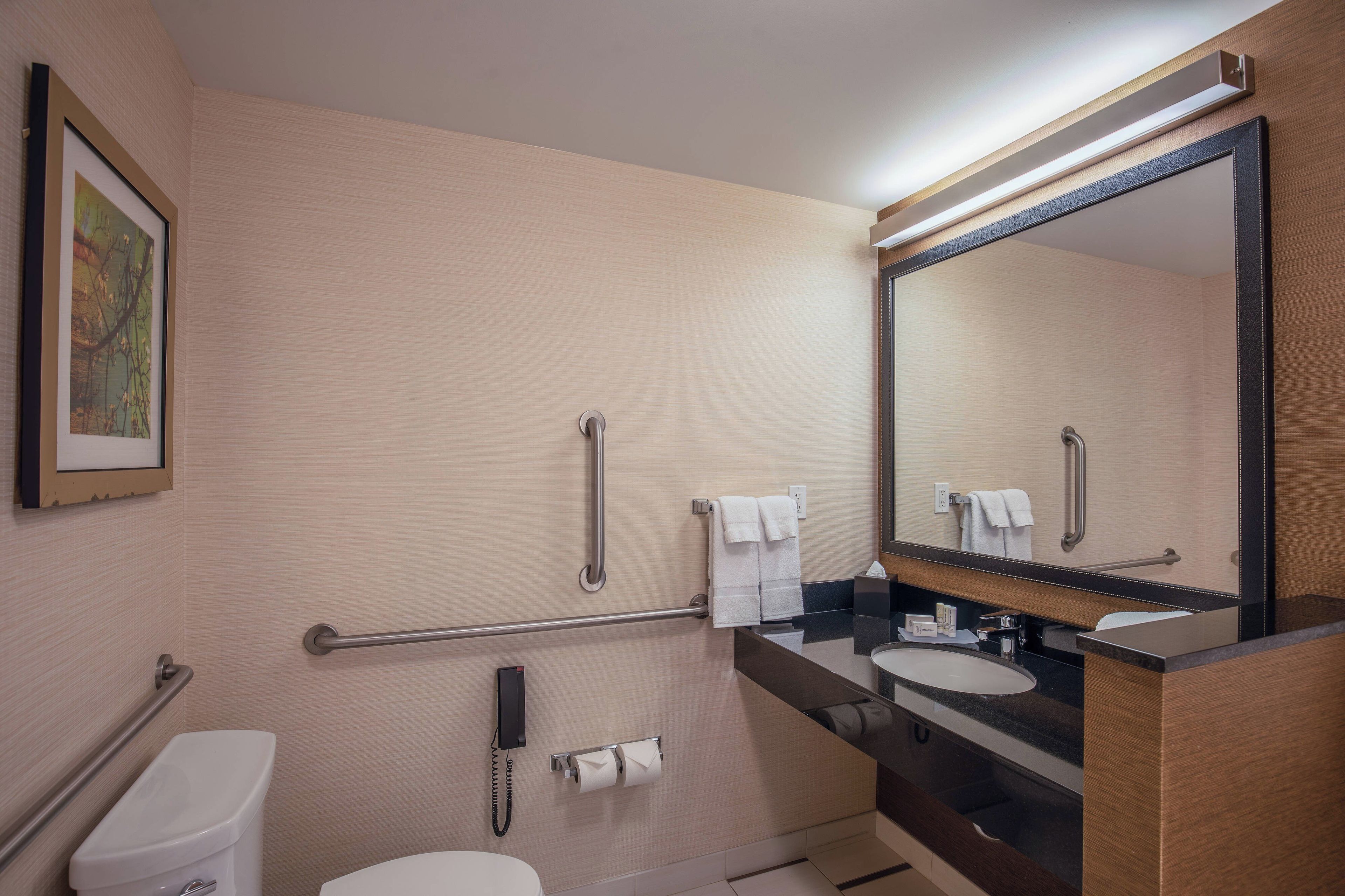 A hotel bathroom with a toilet, bidet, handrails, a sink, a mirror, and a telephone.