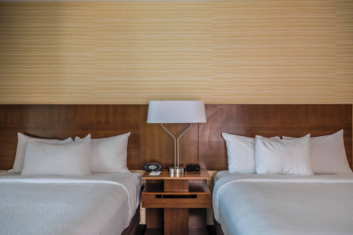 Hotellihuone, jossa on kaksi sänkyä, puinen pääty, yöpöytä lampun kanssa, kello ja pistorasia.