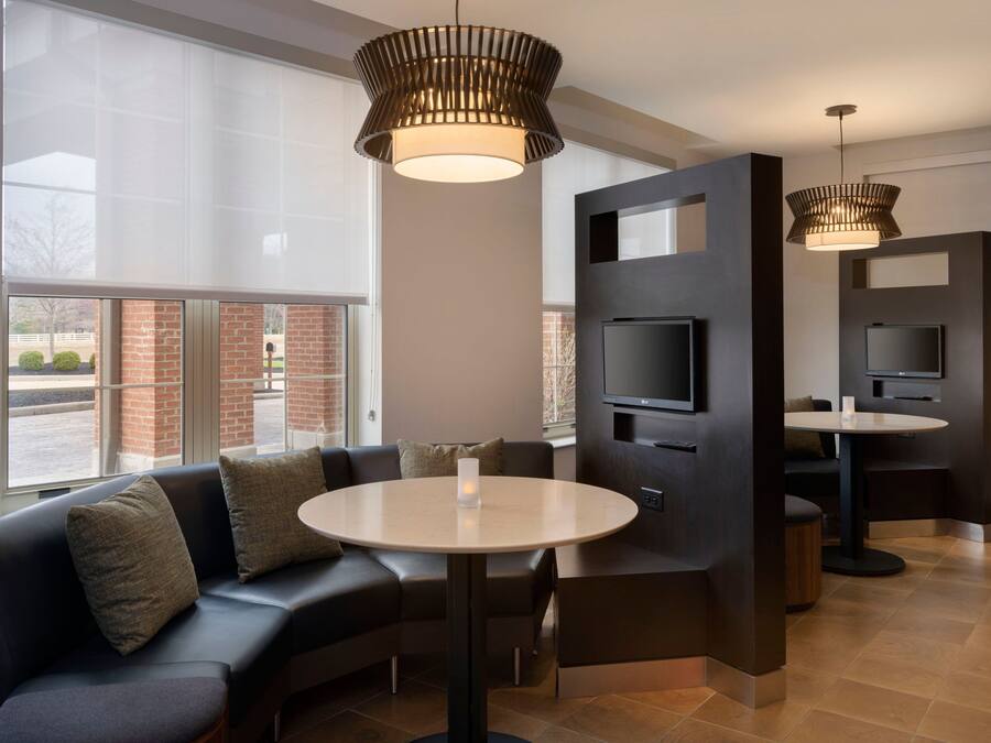 A modern hotel lobby with a sectional sofa, a round table, a flat-screen TV, and a large window offering a view of the outside.