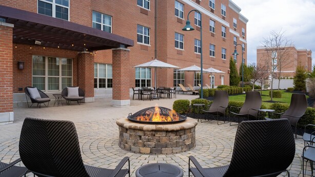 An outdoor patio with a stone fire pit surrounded by black wicker chairs and tables, a covered seating area, and a brick building in the background. - Courtyard Columbus New Albany (New Albany)