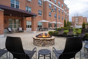 An outdoor patio with a stone fire pit surrounded by black wicker chairs and tables, a covered seating area, and a brick building in the background.