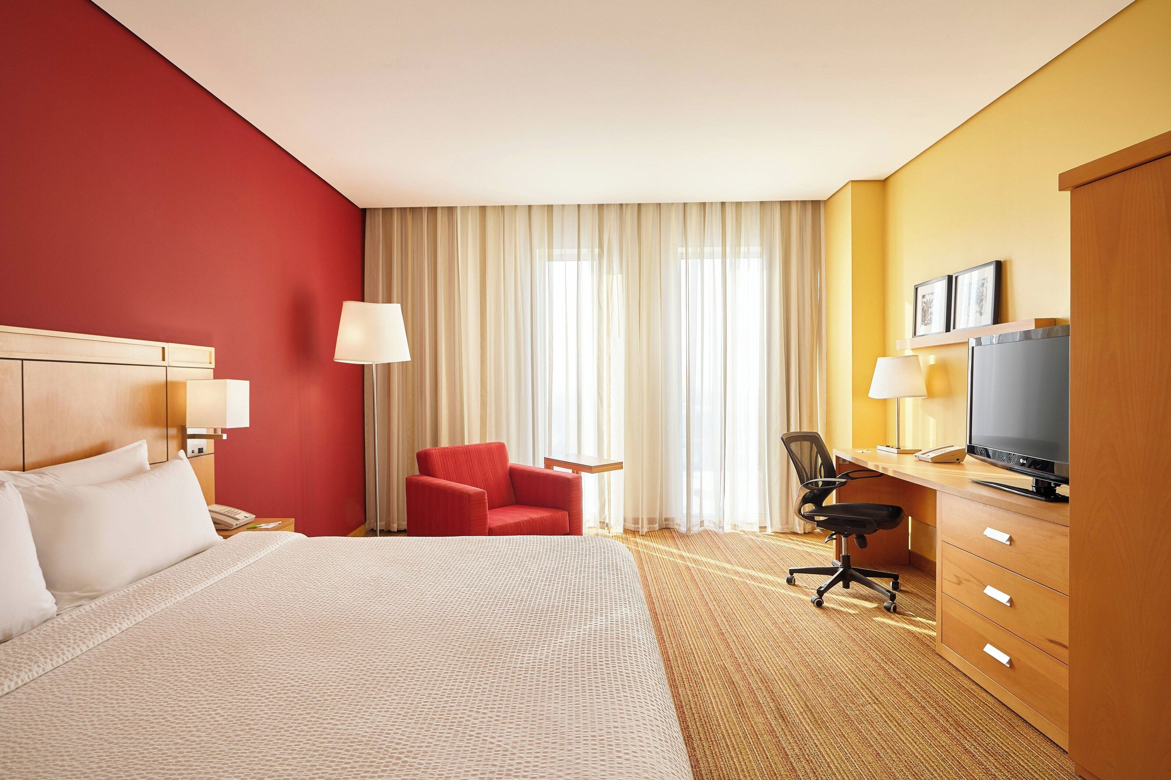 A hotel room with a large bed, a red armchair, a desk with a television, and a window with curtains.