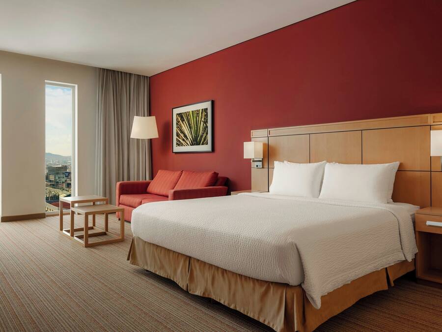 Habitación de hotel con una cama grande, un sillón rojo, una mesita pequeña y una mesita de noche.