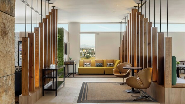 A modern living room with a yellow sofa, a coffee table, and a chair. There is a large window with a view of the city. - Courtyard by Marriott Leon at the Poliforum (León)
