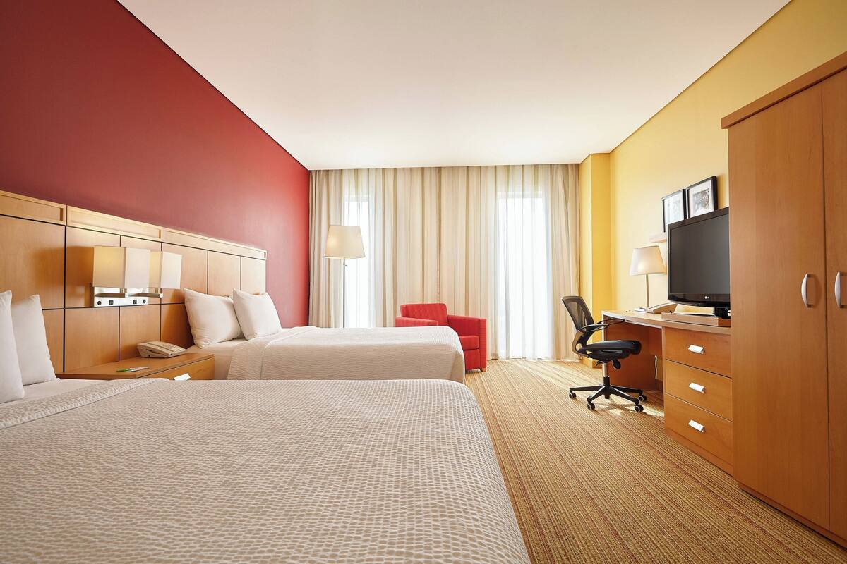 A hotel room with two beds, a desk with a computer, a TV, and a red chair.