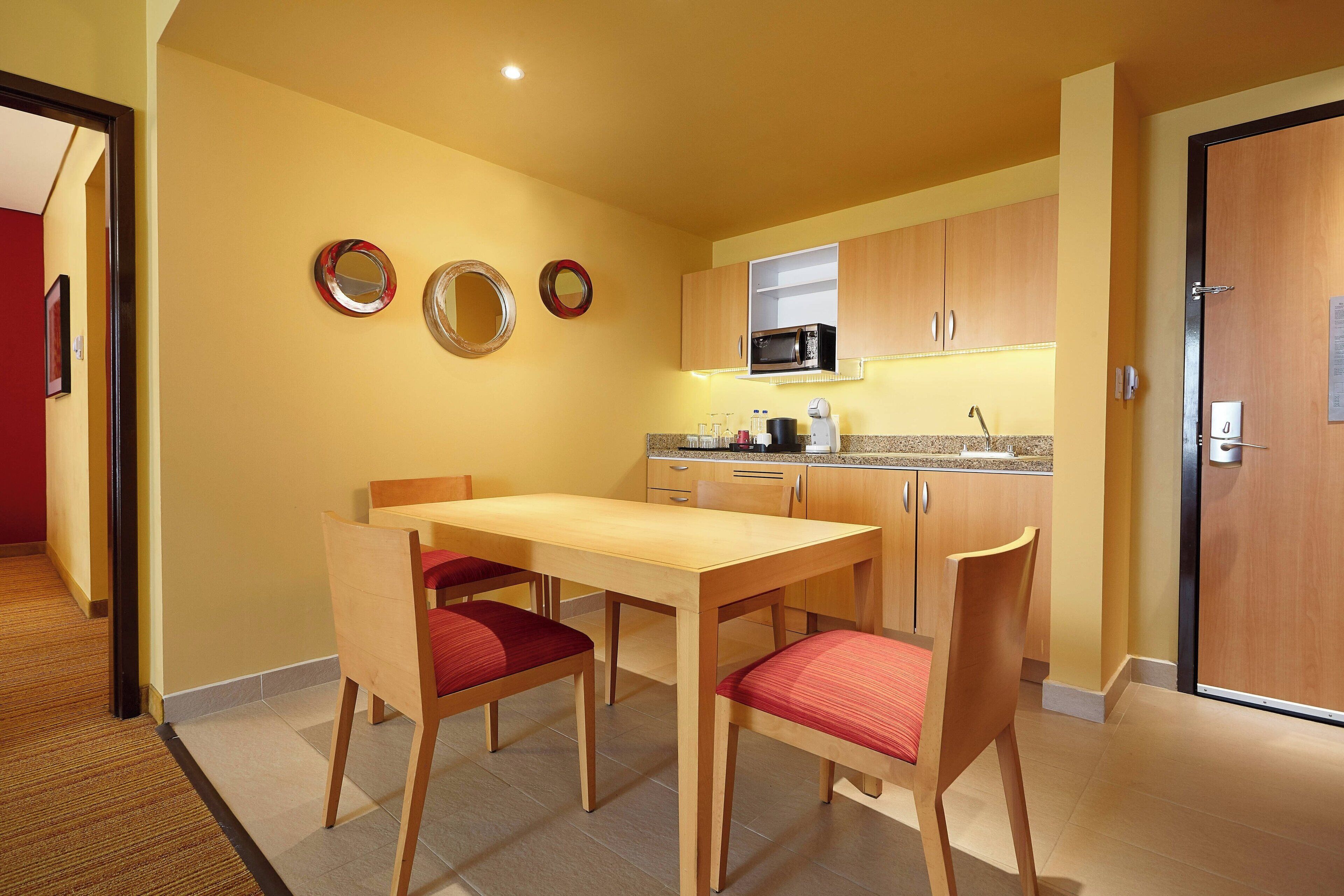 Een kleine keuken met een magnetron, kastjes en een eethoek met stoelen.