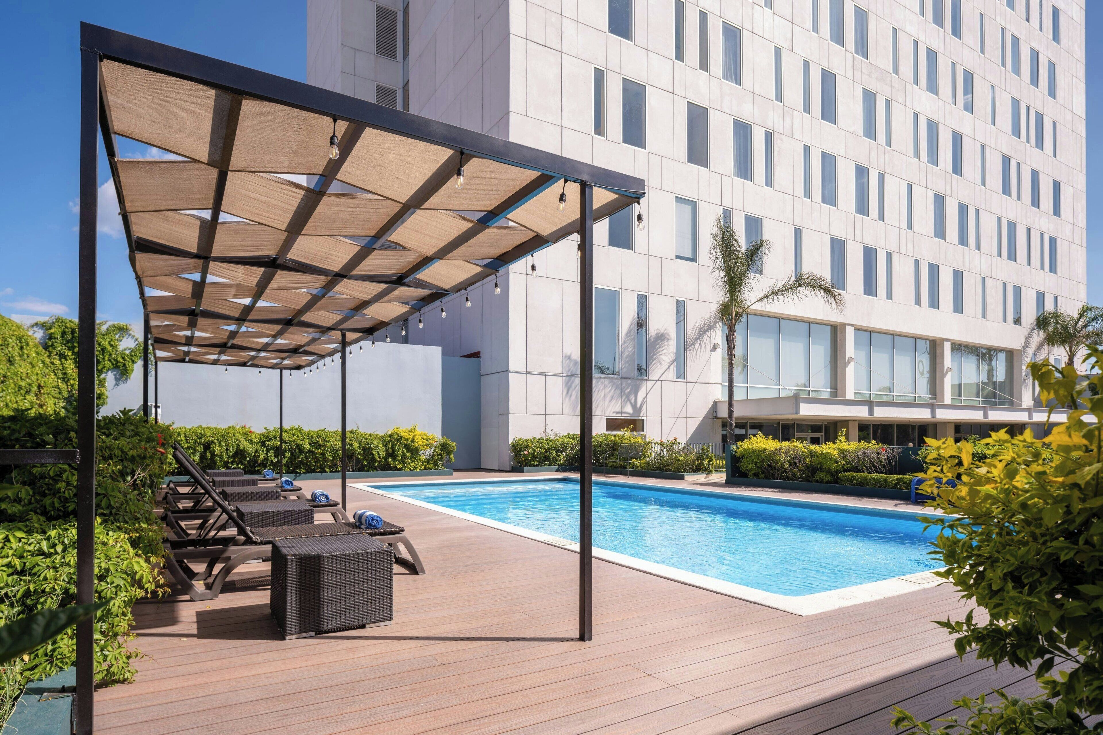Un espace piscine aménagé sur le toit, avec des chaises longues et un auvent, entouré de verdure et bordé d’un bâtiment moderne.