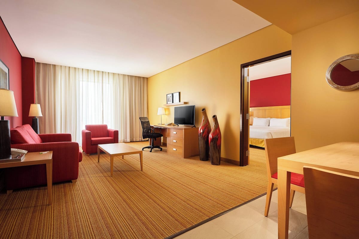 A hotel room with a red sofa, a wooden desk, a television, a bed with a red headboard, and a large mirror on the wall.