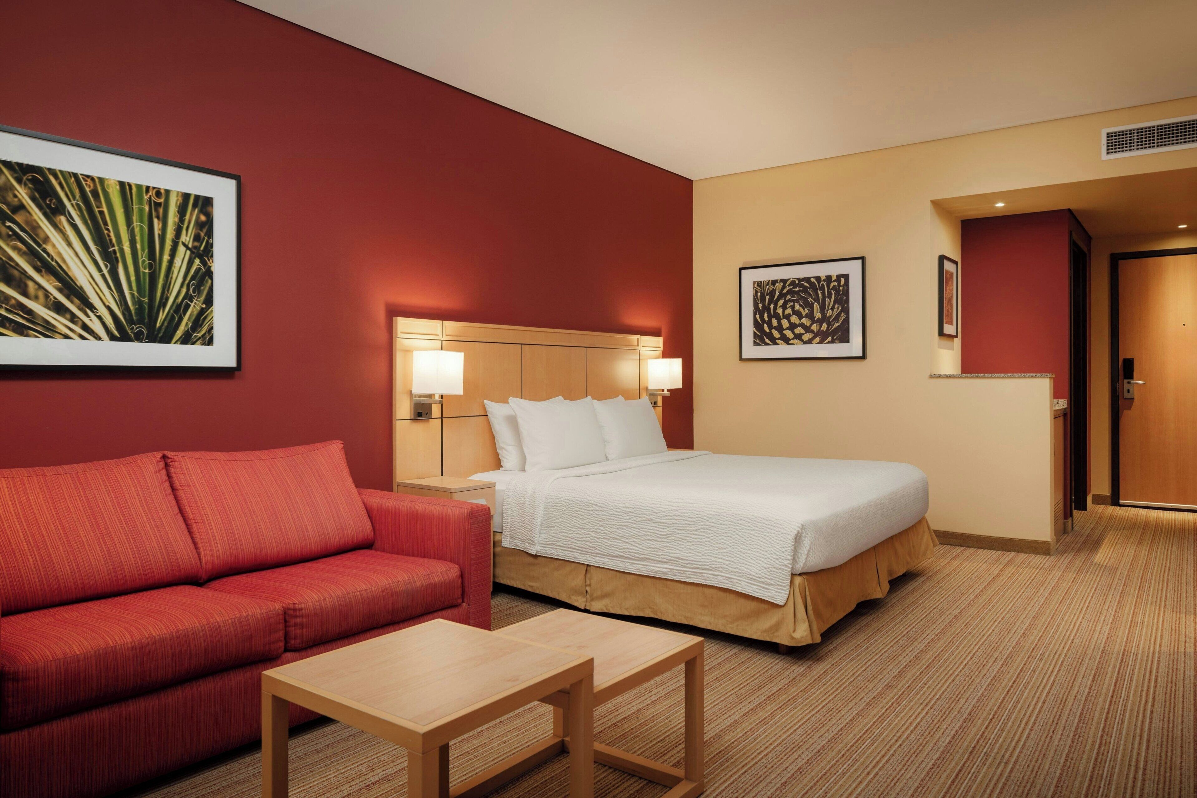 A hotel room with a bed, a red sofa, a wooden table, and two wall-mounted lamps.