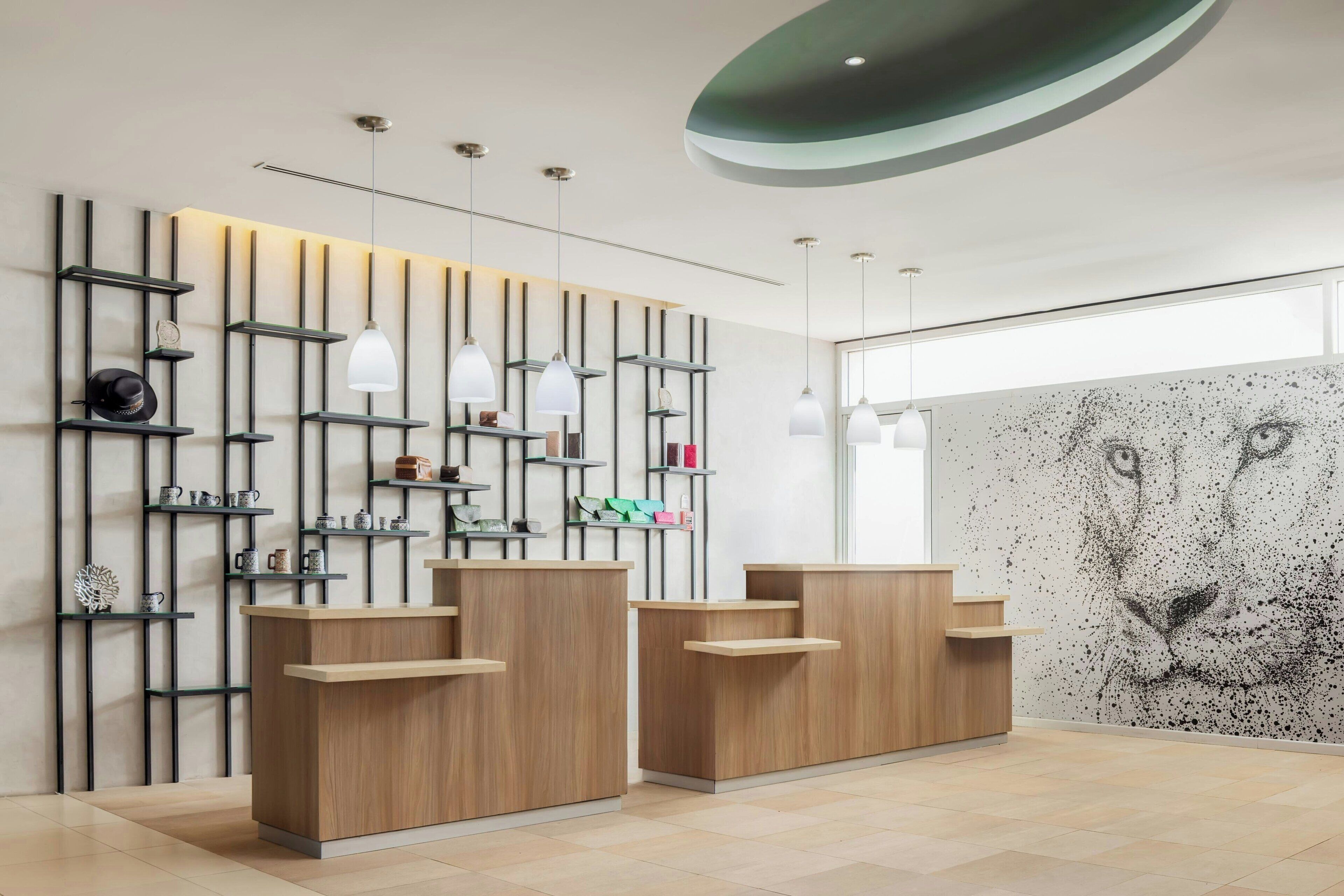 A modern reception area with wooden reception desks, vertical shelving with decorative items, pendant lighting, and a large abstract wall mural.