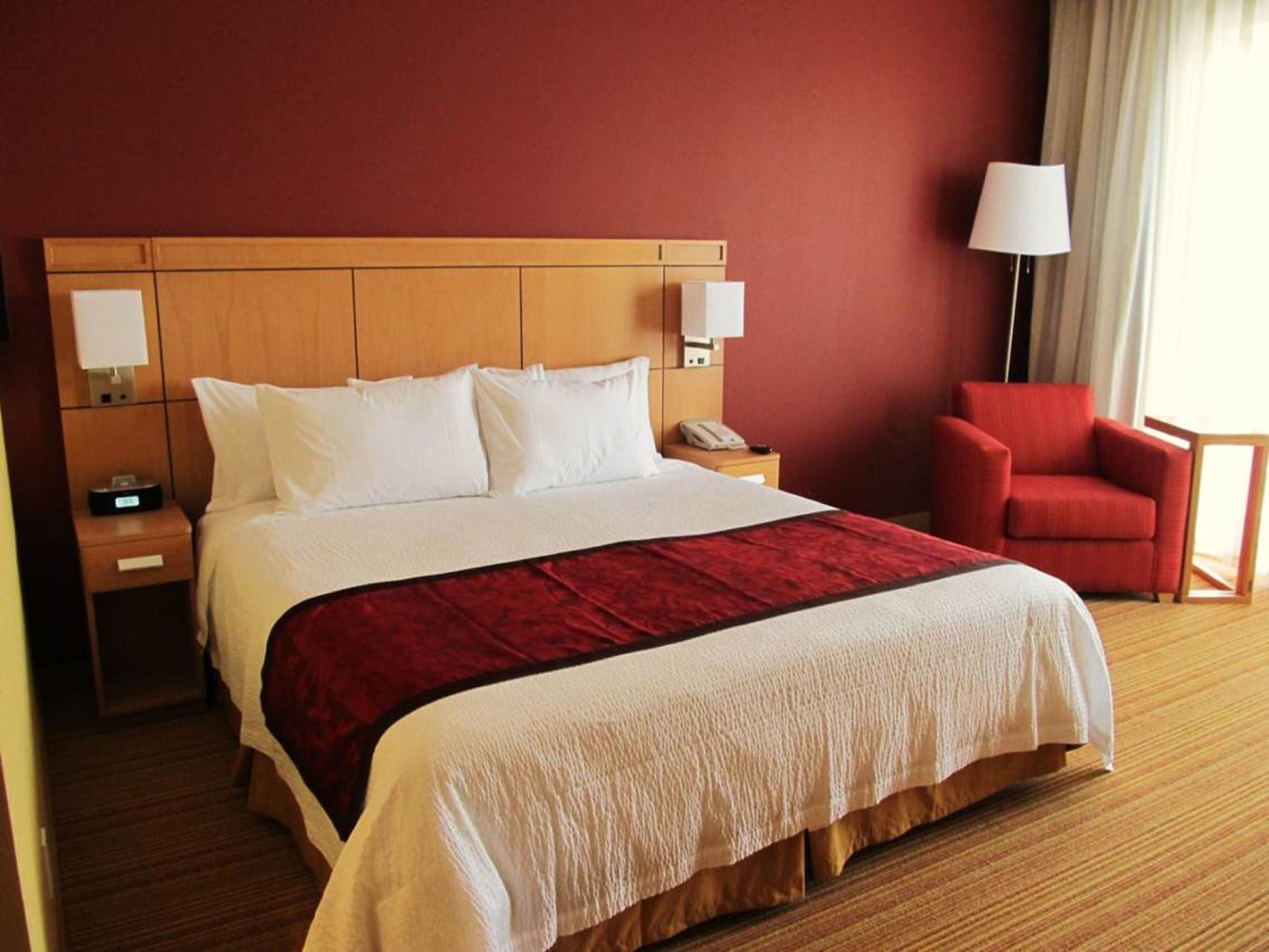 Ein Hotelzimmer mit einem großen Bett, einem hölzernen Kopfteil, einem roten Sessel, einem Nachttisch mit Lampe und einem Telefon.