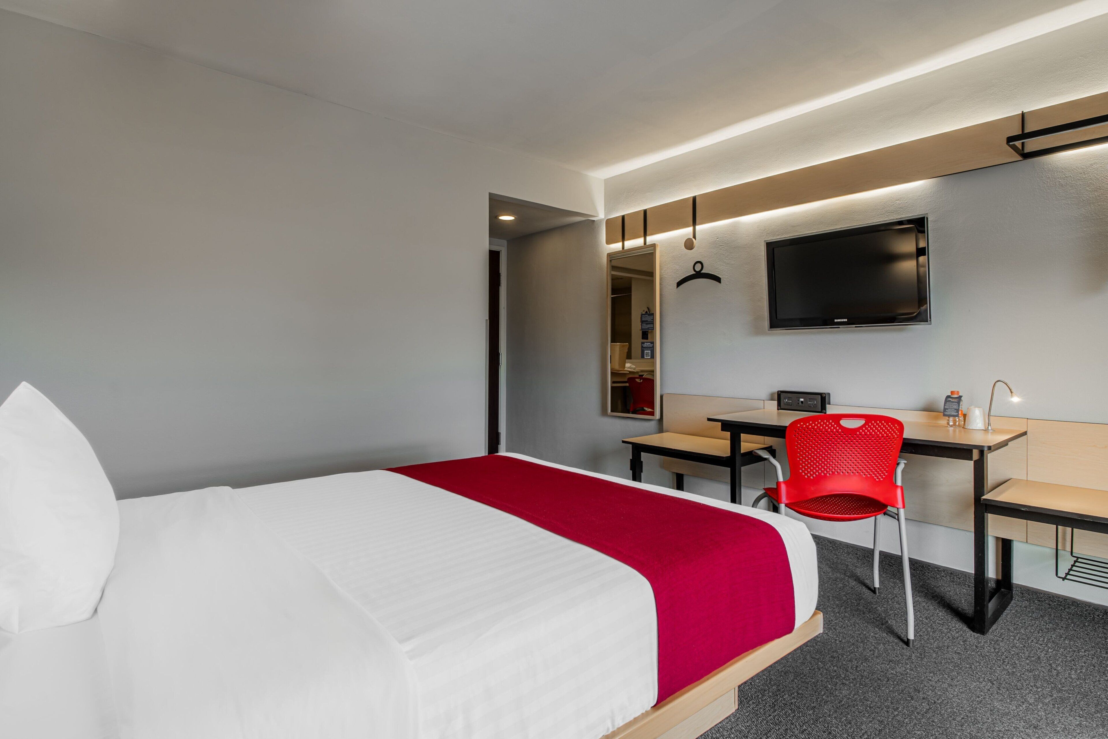Quarto de hotel com cama, escrivaninha, cadeira, televisão e espelho.