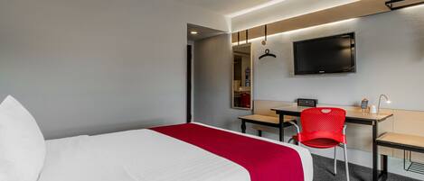Quarto de hotel com cama, escrivaninha, cadeira, televisão e espelho.