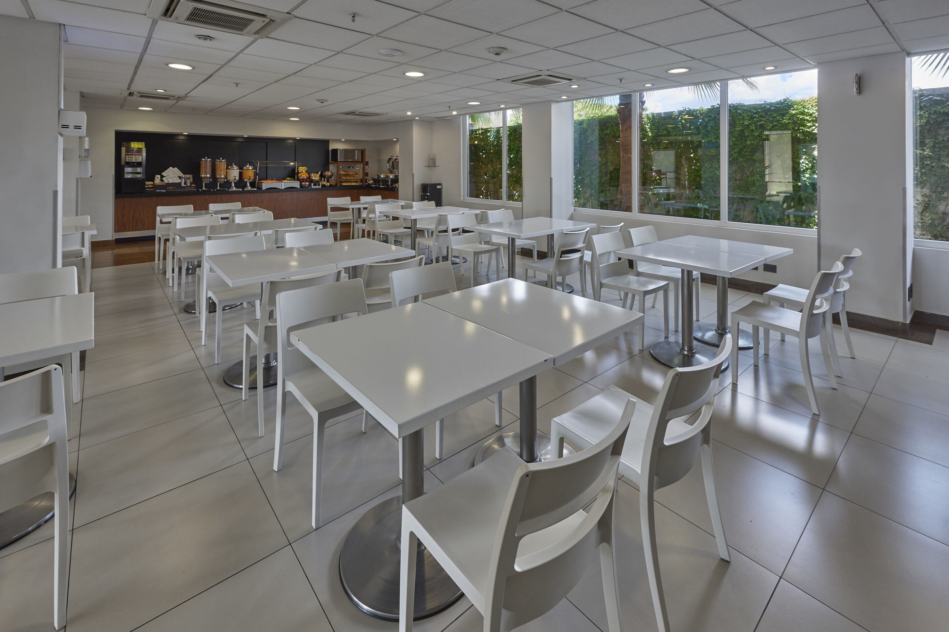 Un comedor amplio con mesas y sillas blancas, una barra con opciones de comida y bebida, y ventanas grandes que ofrecen vistas a zonas verdes.