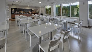 Un comedor amplio con mesas y sillas blancas, una barra con opciones de comida y bebida, y ventanas grandes que ofrecen vistas a zonas verdes.
