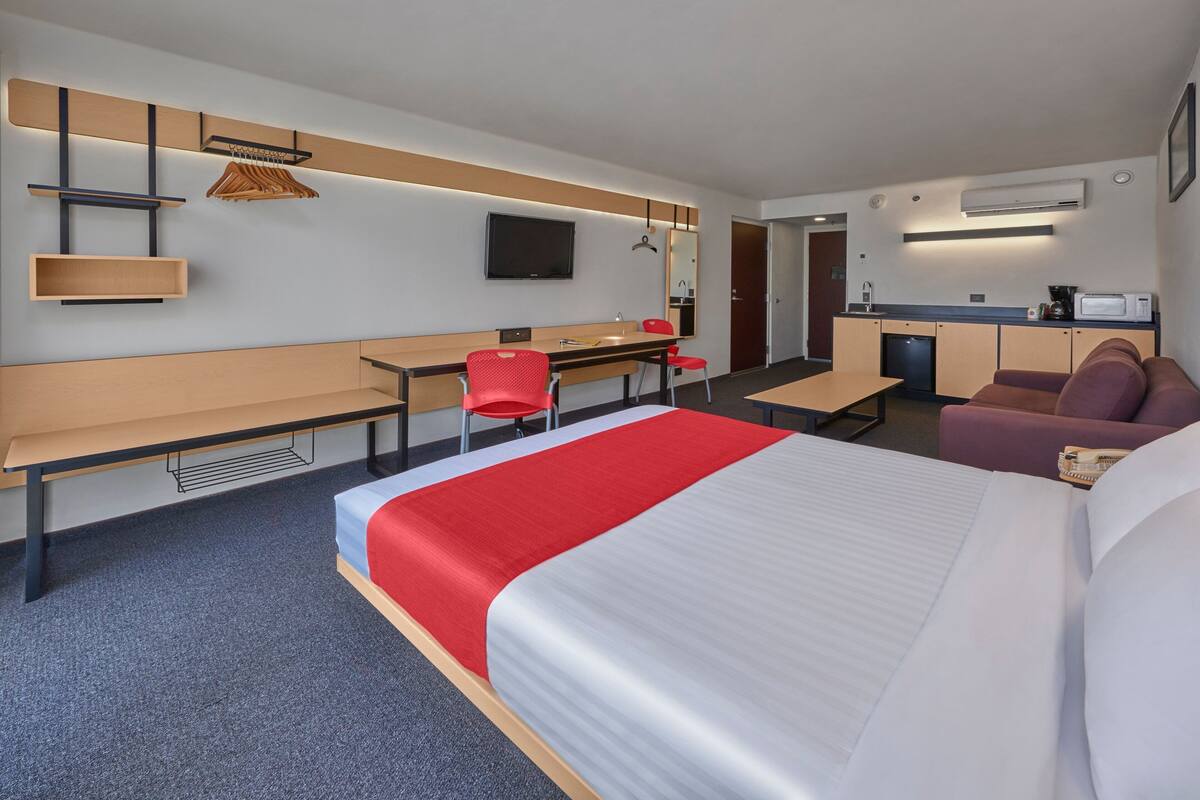 A hotel room with a bed, a desk, a chair, a sofa, a TV, a mirror, and a kitchenette.