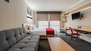 A modern hotel room with a grey sofa, a bed with a red accent, a desk with a chair, and a TV mounted on the wall. - City Express by Marriott Querétaro Juríca (Querétaro)