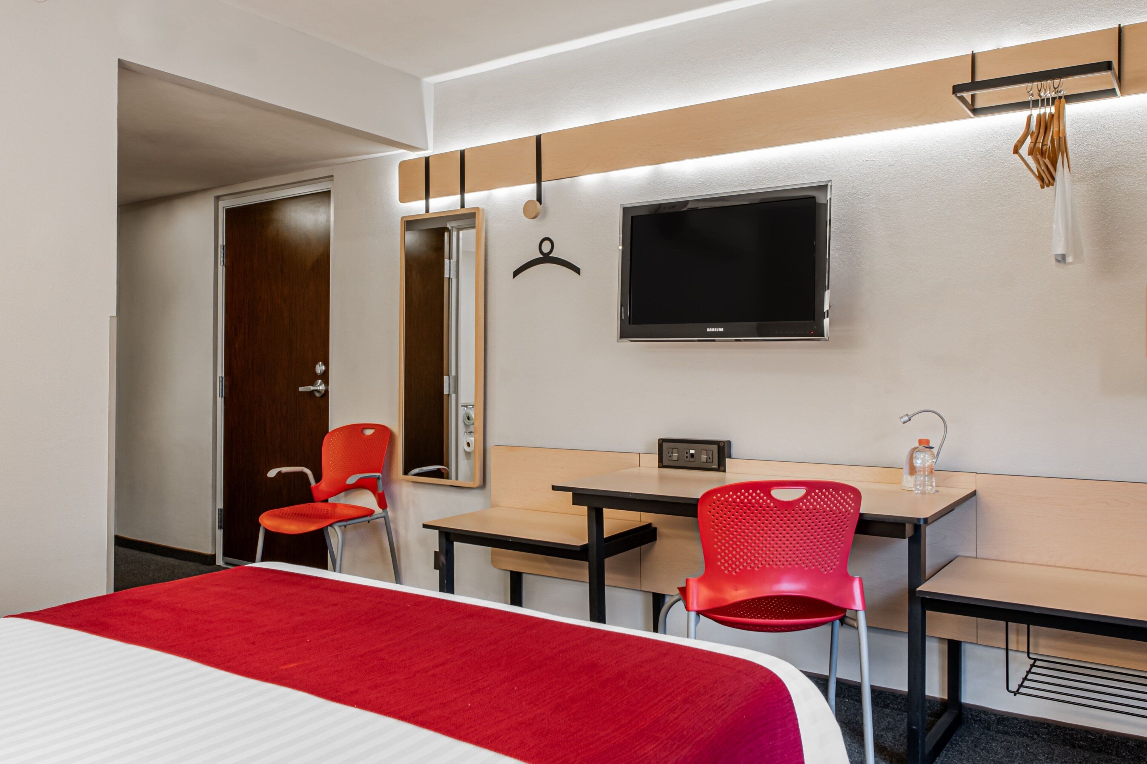 Una habitación de hotel con una cama, dos sillas rojas, un escritorio, un televisor y un espejo.