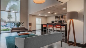 A modern hotel lobby with a grey sofa, a wooden coffee table, a floor lamp, and a bar area with red chairs. - City Express by Marriott Querétaro Juríca (Querétaro)