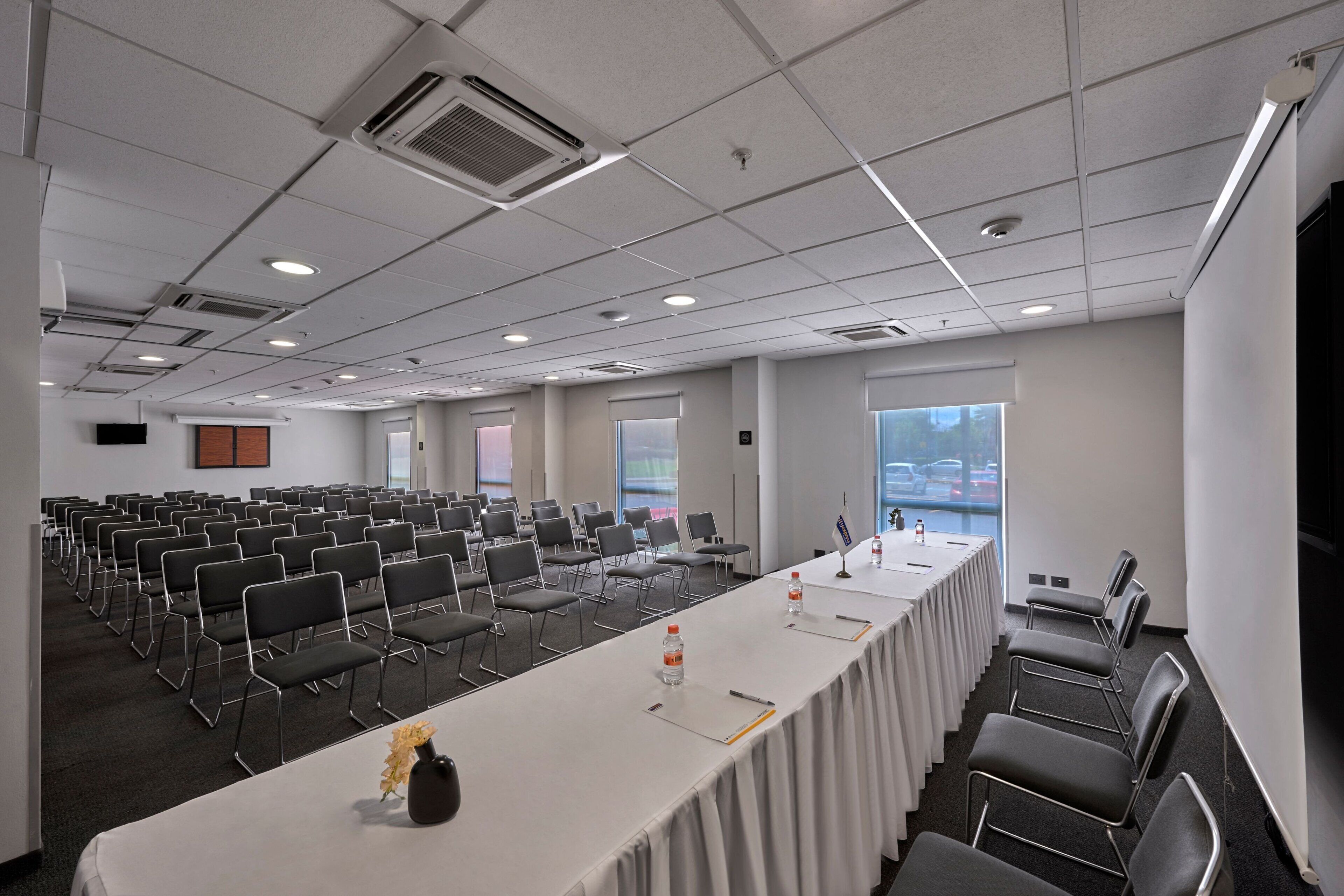 A conference room with rows of chairs, a long table with water bottles, and a projector screen.