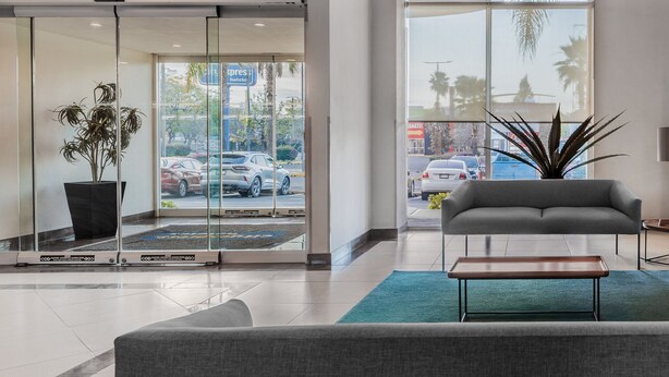A modern hotel lobby with a sliding glass door entrance, a grey sofa, a wooden coffee table, and a large potted plant. - City Express by Marriott Querétaro Juríca (Querétaro)