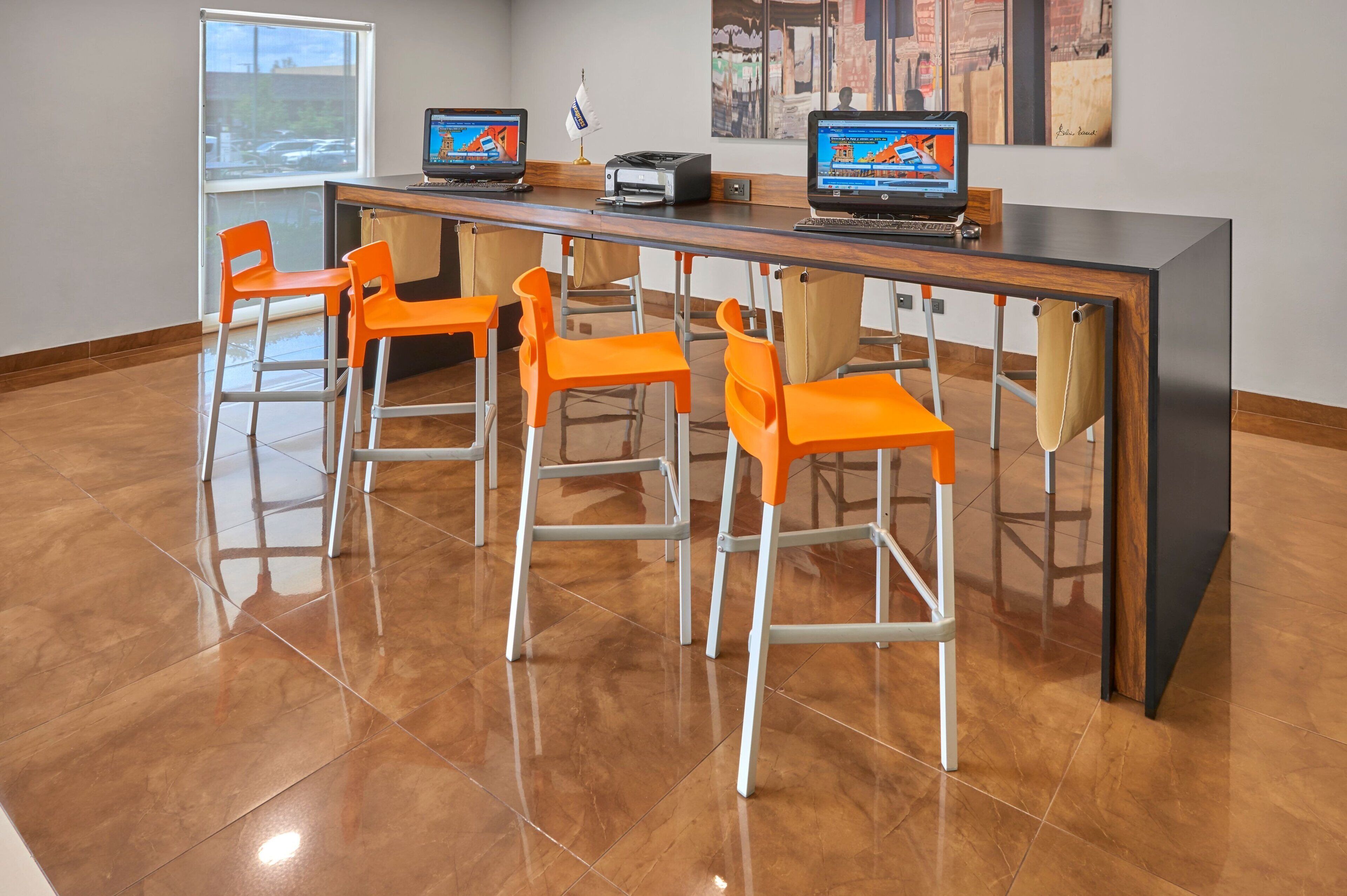 Zona de bar moderna con una barra alargada, varias sillas color naranja y dos computadoras portátiles sobre la barra.