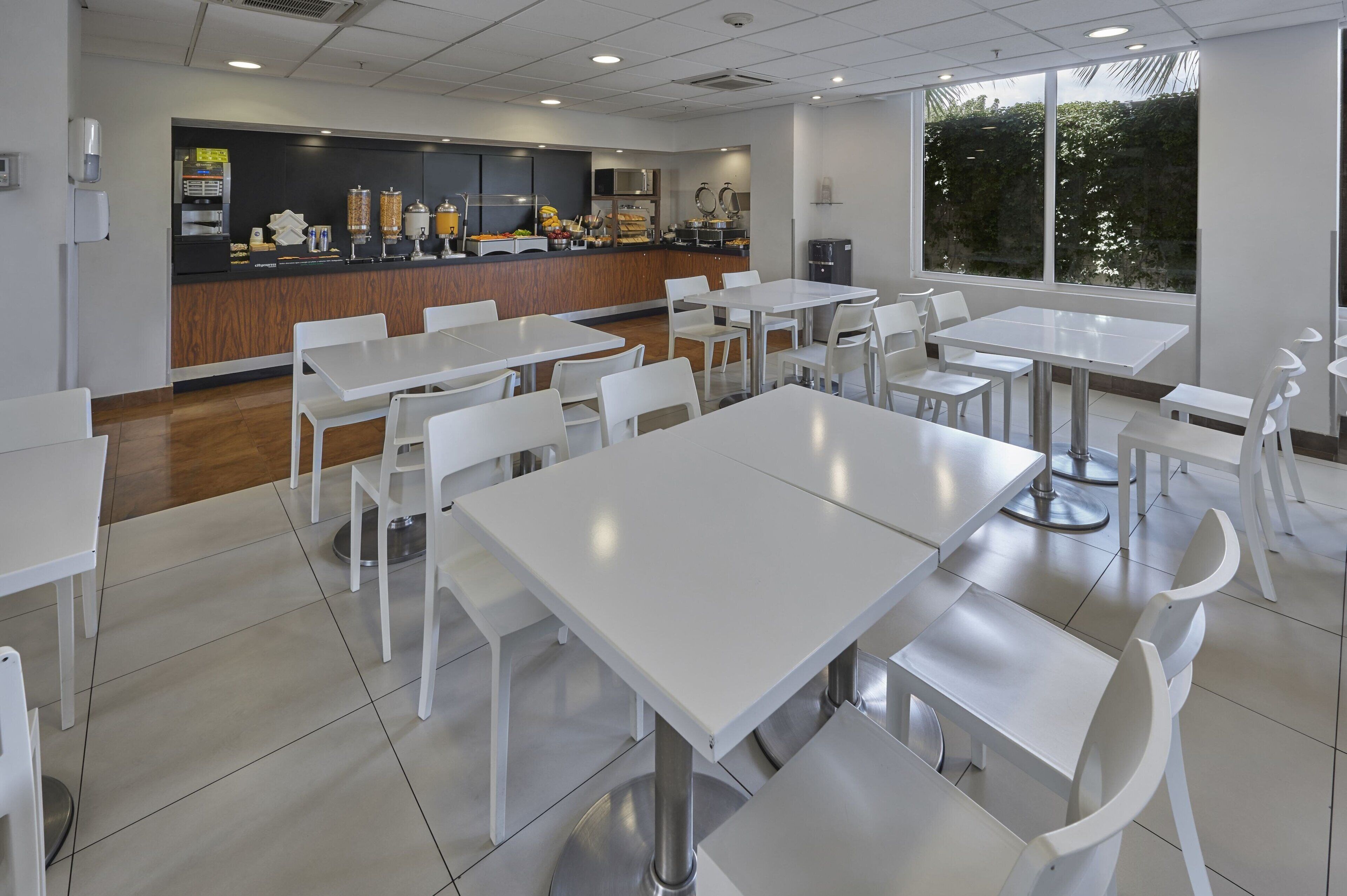 A modern dining area with white tables and chairs, a counter with food items, and a view of greenery outside.
