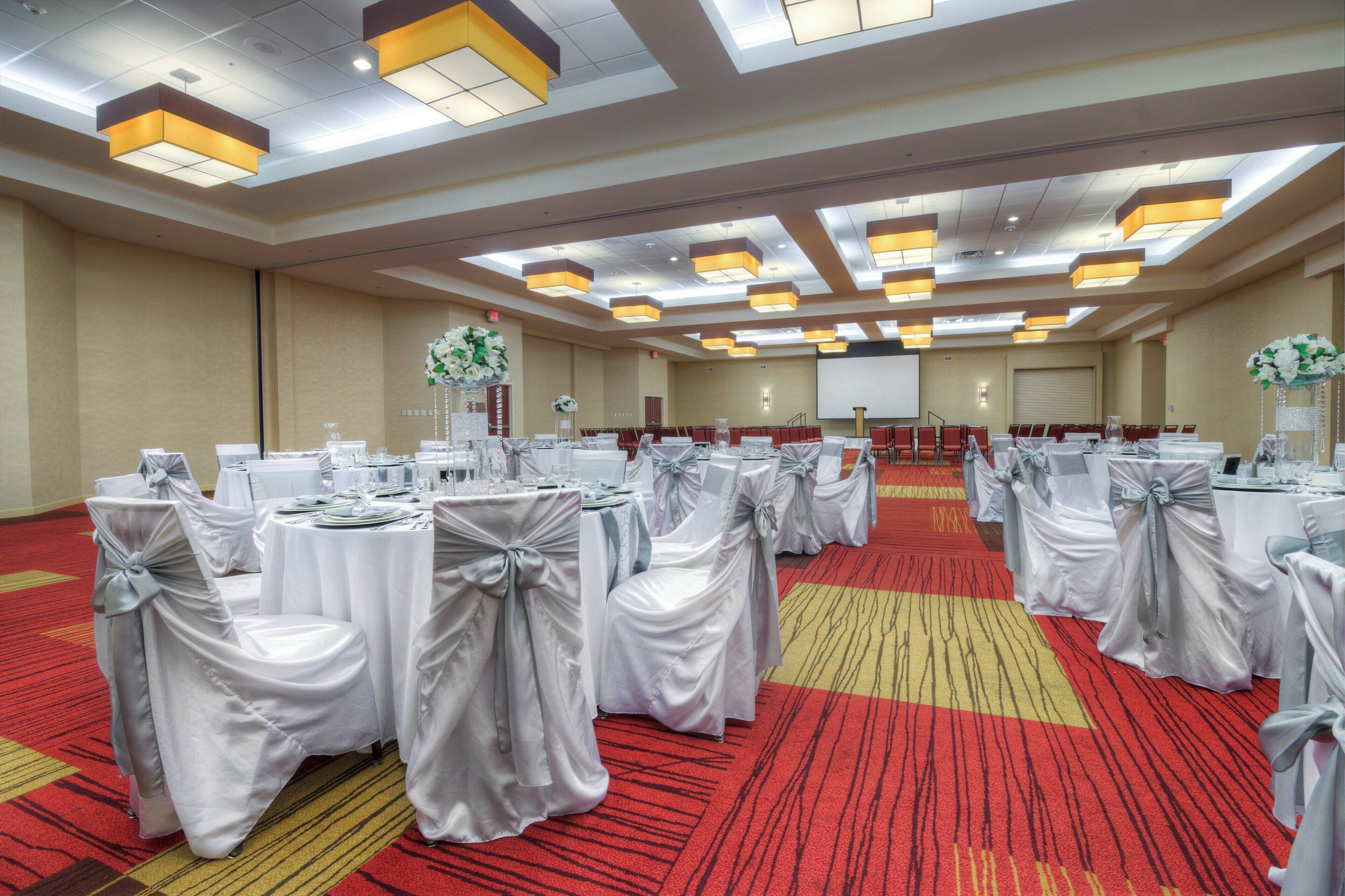 A conference room with round tables, each covered with white tablecloths and silver sashes, and chairs with white covers and silver sashes.