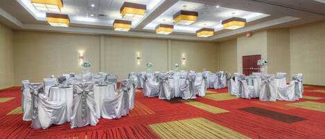 Un salón de conferencias amplio con varias mesas redondas cubiertas de manteles blancos y fajas plateadas, dispuestas en filas.