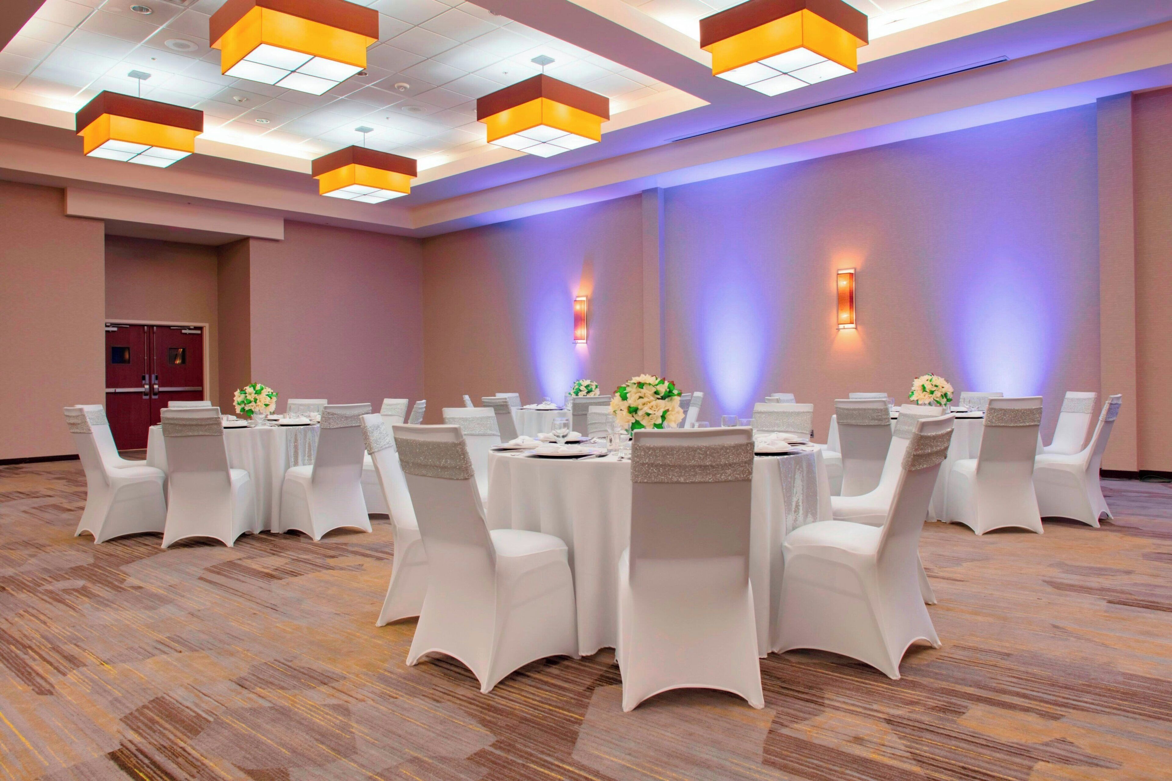 Uma sala de conferências com mesas redondas e cadeiras brancas, equipada com luminárias de teto e arandelas.