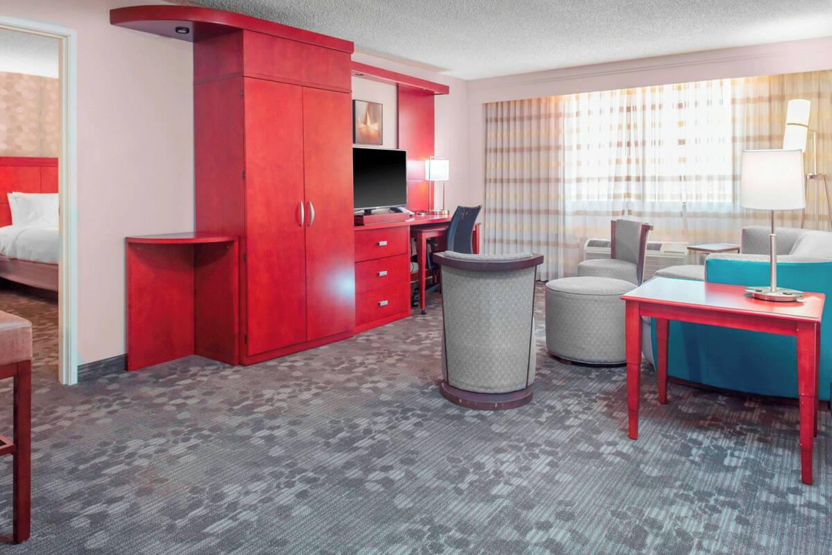 A hotel room with a red wardrobe, a desk with a computer, a chair, a round ottoman, and a small table with a lamp.