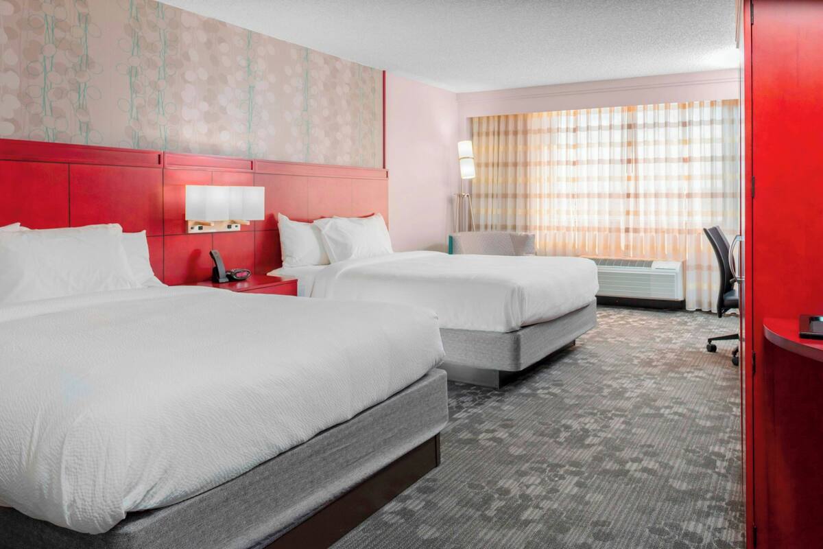 A hotel room with two beds, a red headboard, a desk with a lamp, and a large window with curtains.