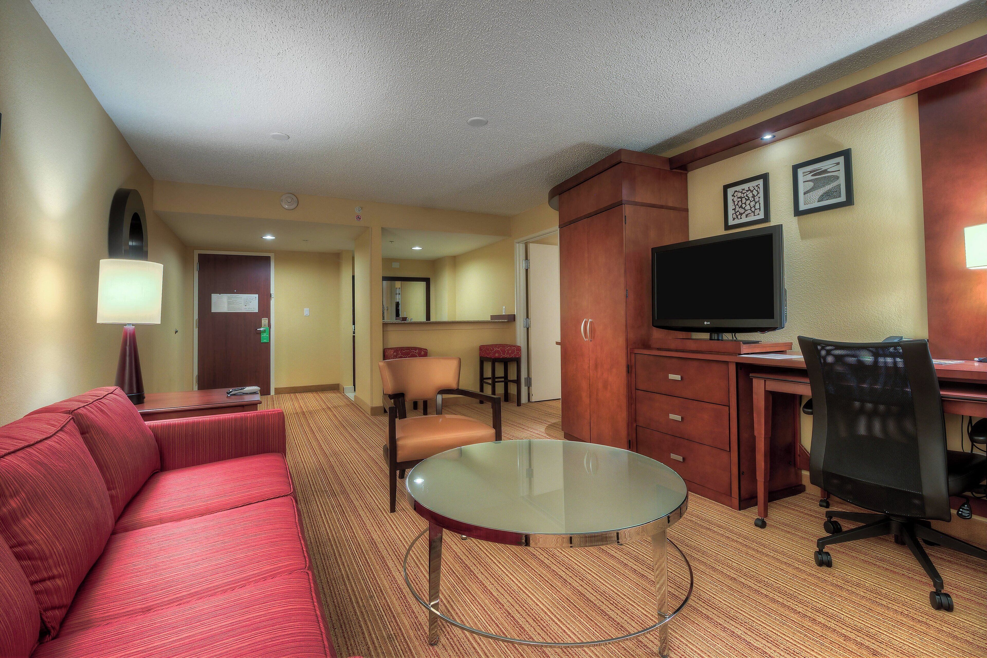 A hotel room with a red sofa, a glass table, a TV, a desk, and a chair.