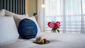 Cama bem arrumada com lençóis brancos, almofada azul com texto, uma xícara de café e uma bandeja com macarons.