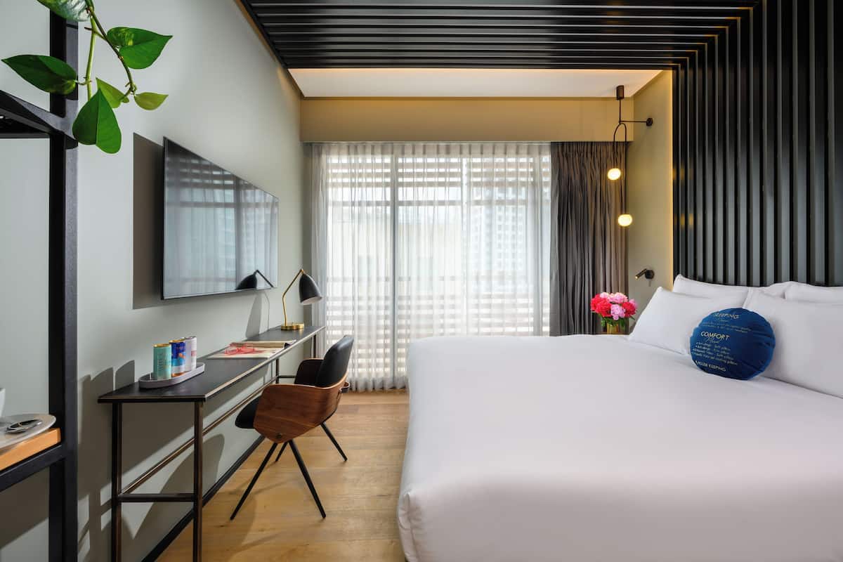 Une chambre d’hôtel moderne, dotée d’un grand lit, d’un bureau avec une chaise, d’une télévision et d’une fenêtre avec des rideaux légers.