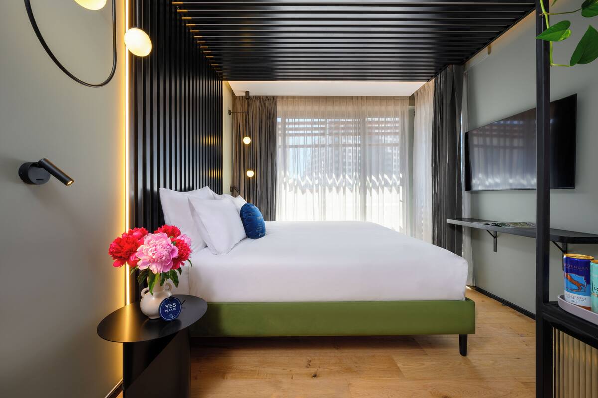 Une chambre d’hôtel moderne avec un lit vert, du linge de lit blanc et une petite table de chevet agrémentée d’un vase de fleurs.