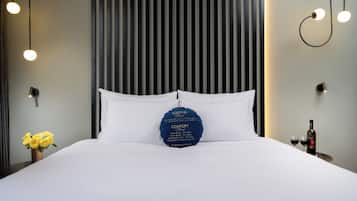 A neatly made bed with white linens, a blue pillow with text, and a bottle of wine on a bedside table.