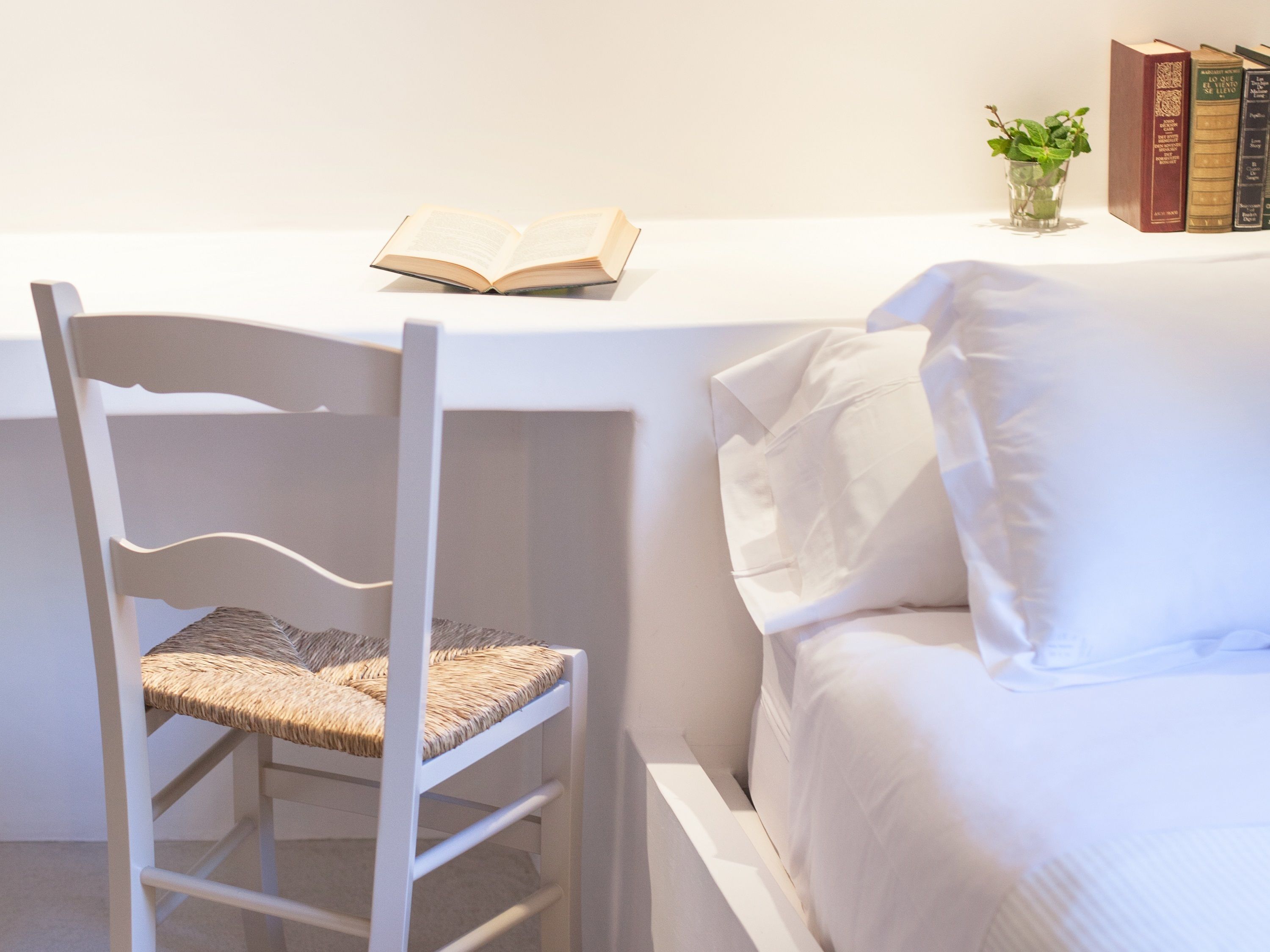 A white chair with a woven seat, a white desk with an open book, and a white bed with pillows.