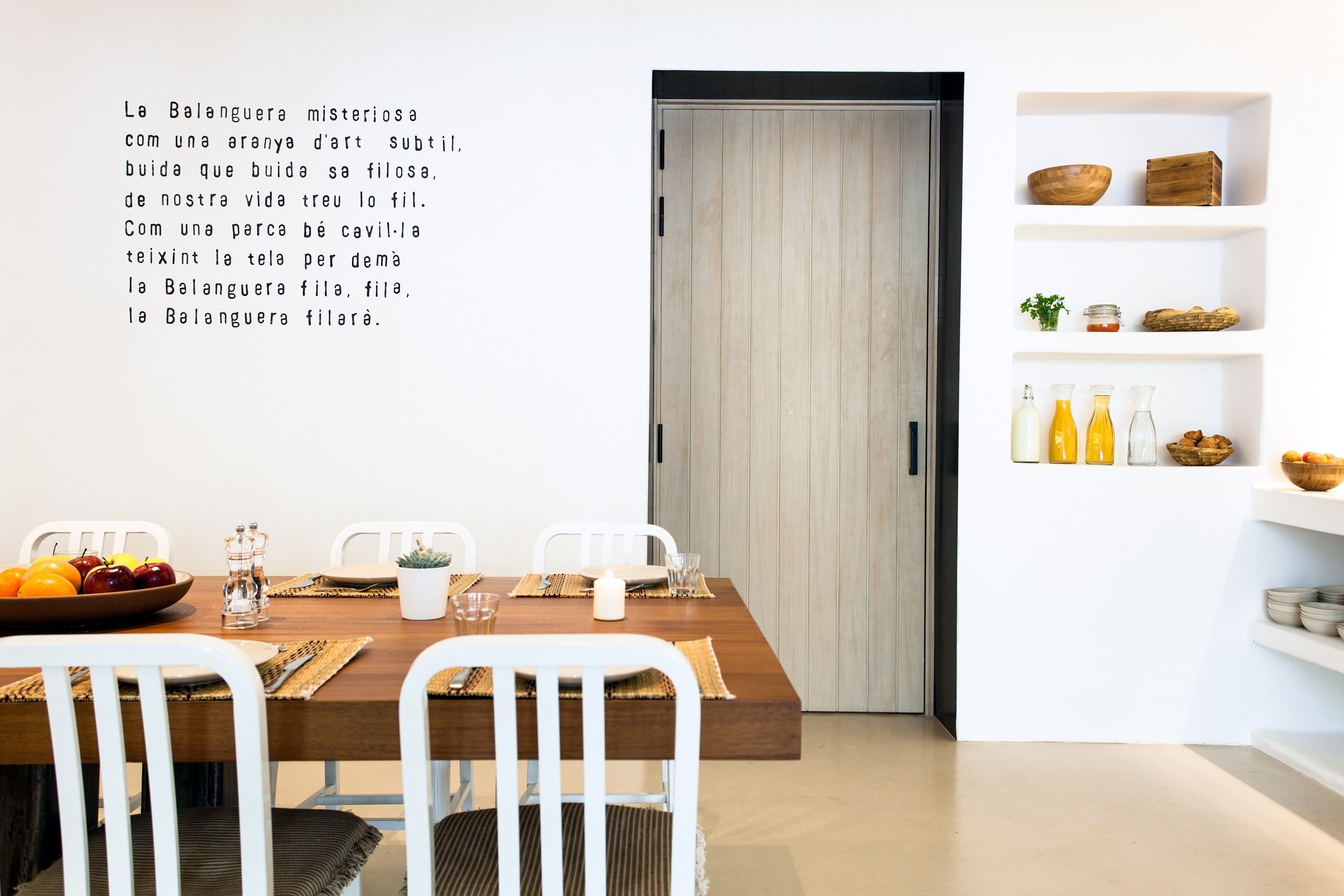 Un comedor moderno con una mesa de madera dispuesta para cuatro, sillas blancas y una pared blanca con estantes que contienen utensilios de cocina.