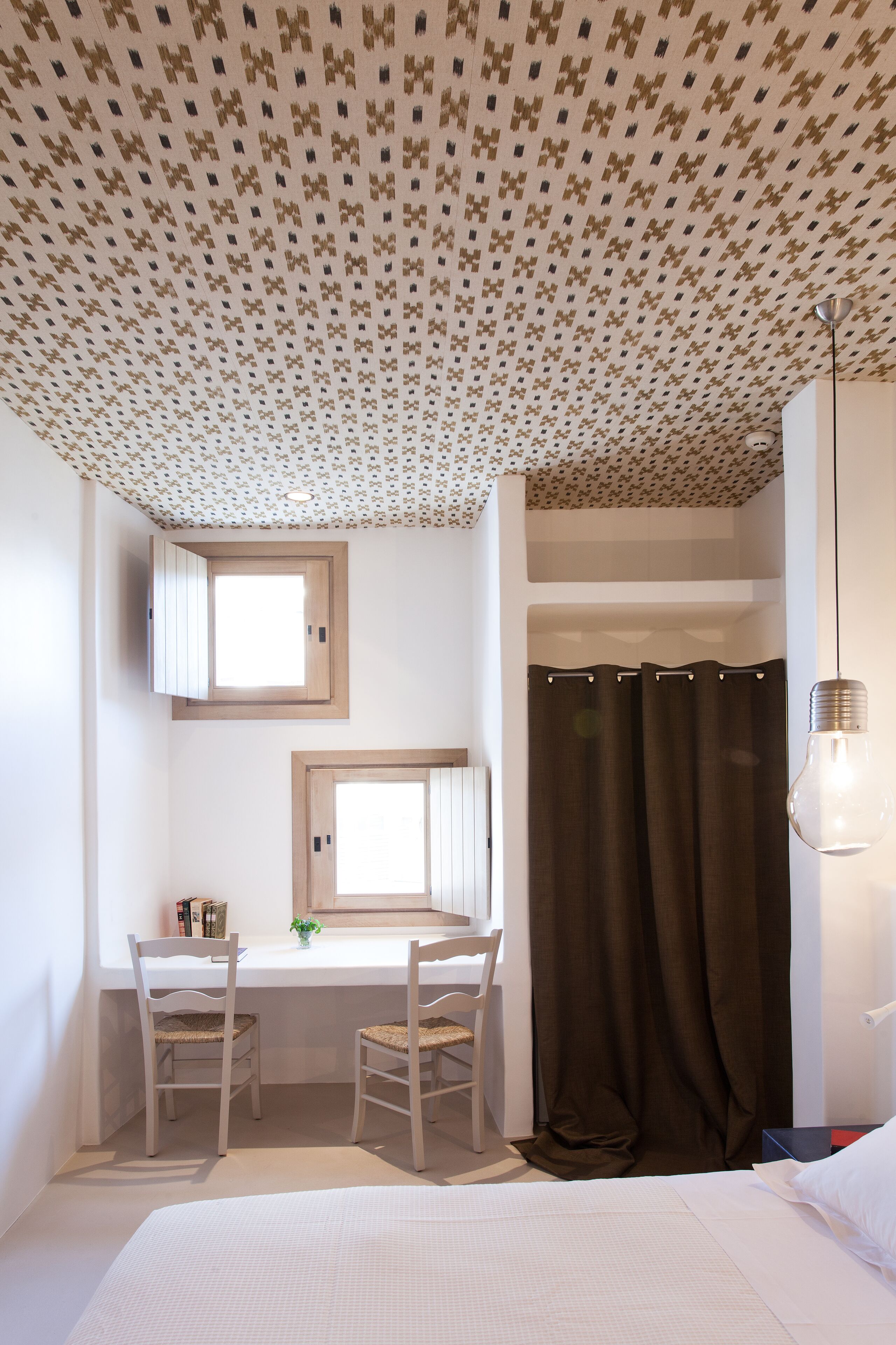 Un dormitorio con cama, escritorio, dos sillas, ventana, cortina e iluminación colgante.