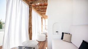 A modern, well-lit room with a wooden ceiling, a white sofa with black and beige pillows, a round white ottoman, and a small table with a white candle.