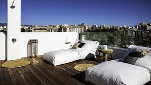 A rooftop terrace with two white beds, a small table, and a wall-mounted light fixture.