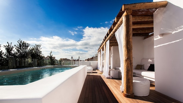 A rooftop pool area with a wooden deck, white walls, and a covered seating area with cushions.