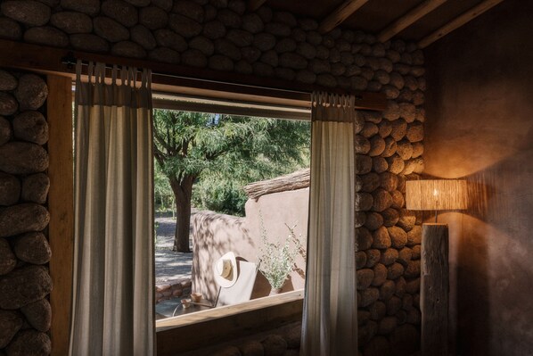 A stone wall with a wooden beam ceiling, a window with curtains, and a lamp.