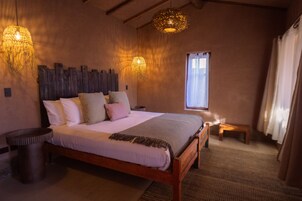 Un dormitorio con una cama de madera, dos lámparas de noche, un ventanal con cortinas y un taburete de madera.