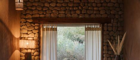 A bedroom with a stone wall, a bed, a nightstand, a lamp, and a view of the outdoors.