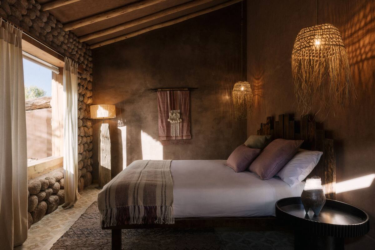 A bedroom with a bed, bedside table, lamp, and a view of the outside.