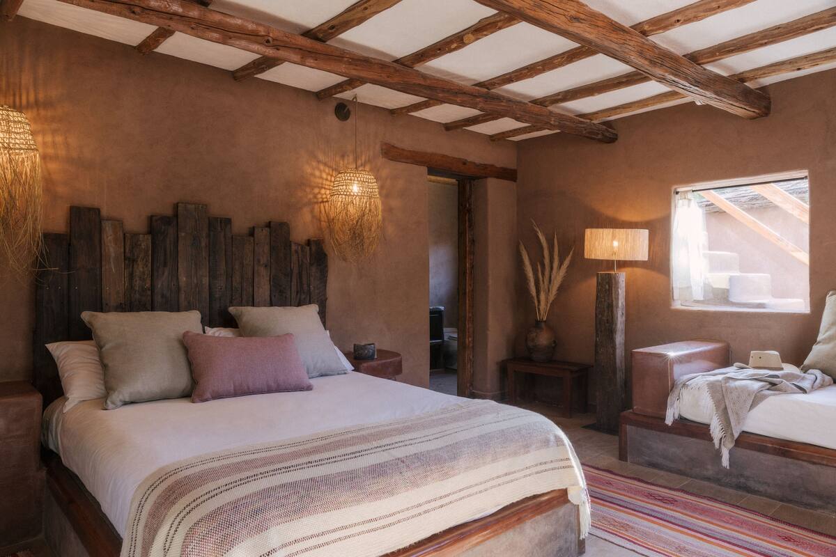 A bedroom with a large bed, wooden beams on the ceiling, a bedside lamp, and a window with a view.
